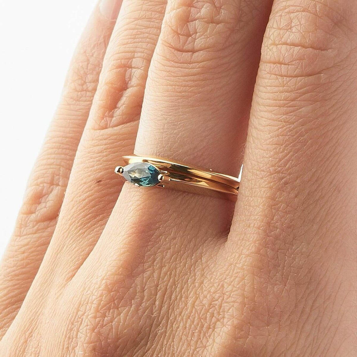 A close-up of a hand wearing a yellow gold ring set with a marquise-cut teal Australian sapphire.