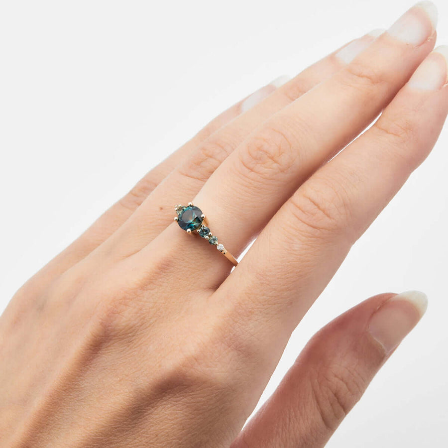 A hand displaying a yellow gold ring with an Australian sapphire at the center, complemented by diamonds and a square band design.