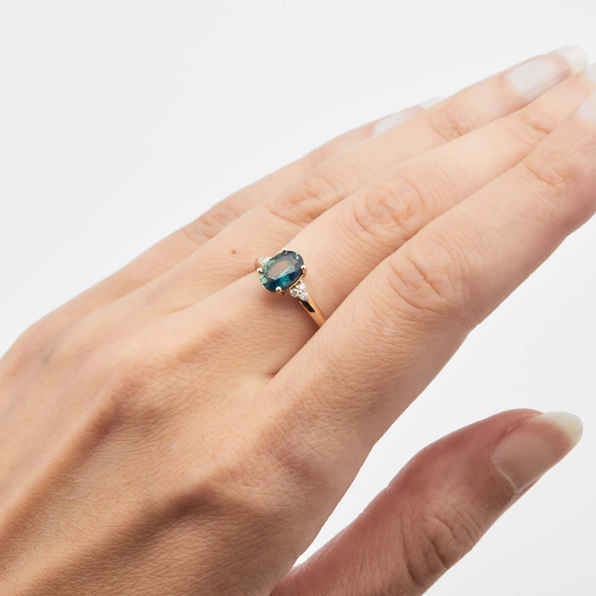 A hand wearing a yellow gold ring with a brilliant cut teal Australian sapphire in a triangle diamond arrangement on a knife-edge band.