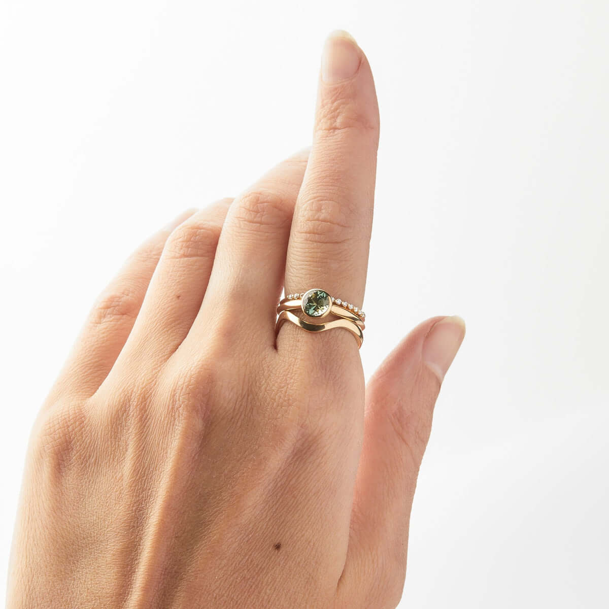 A hand model displaying 3 gold rings. The 1st a thin band covered in small diamonds. The 2nd a large green sapphire and 3rd a wavy gold band. 
